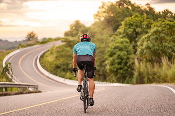 back view of a cyclist on top of a mountains winding road, riding a black bicycle down a hill, wearing bike helmet and blue cycling jersey, with grey clouds sunset sky and forest in the background.