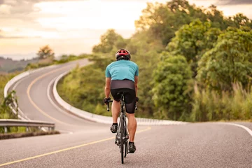 Fotobehang back view of a cyclist on top of a mountains winding road, riding a black bicycle down a hill, wearing bike helmet and blue cycling jersey, with grey clouds sunset sky and forest in the background. © Have a nice day 