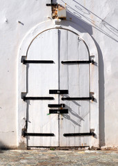 Weathered white wooden double arched doors with black hinges set into a white wall with white trim along the cobblestone sidewalks of old town St. Thomas U.S. Virgin Islands.