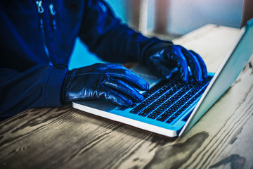 closeup concept of a malicious hacker using laptop computer typing and writing code of computer viruses, wearing black hoodie jacket and black leather gloves with a mask, on n wooden computer desk