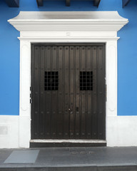 A heavy black metal double door with wrought iron covered widows framed into a vibrant blue wall surrounded by white panted wood trim on a black brick sidewalk.