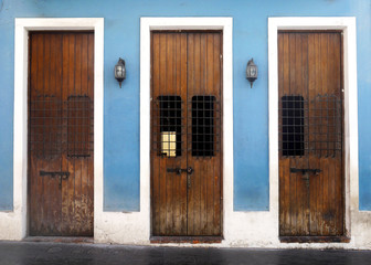 Three old weathered wooden doors with wrought iron covered widows framed into a vibrant blue wall and surrounded by weathered white trim on a black brick sidewalk. - obrazy, fototapety, plakaty