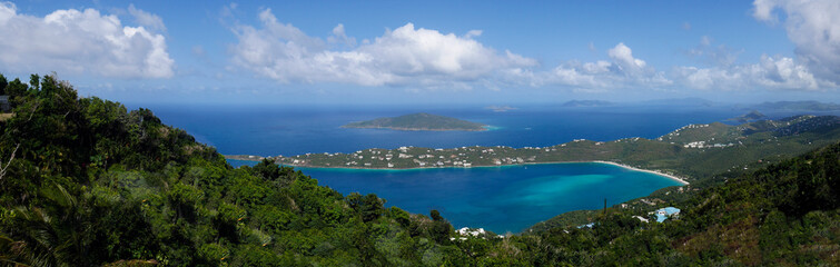 Panoramic view of a small bay on a remote Caribbean island with lush trees , aqua water and a blue sky with white clouds.