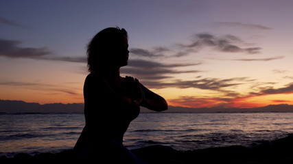 Silhouette of girl sits in namaste pose meditating by the sea at sunset.