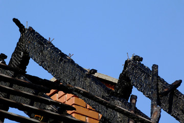 Fototapeta na wymiar charred and burnt black timber roof structure after house fire. abstract view of rafters. blue sky. fire safety and insurance concept. fire hazard, flammable materials in construction. brick chimney.