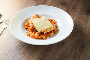 Pasta, Italian food, with tomato sauce topping with deep fried crispy pork cutlet and a sheet of cheese in white dish place on dark brown wooden table and stainless steel utensil near it