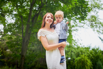 Beautiful young woman in a white dress and long hair with a cute little baby blonde boy at the green garden in a day