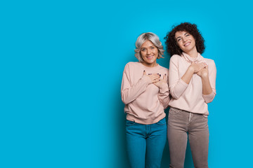 Two delight caucasian sisters with curly hair are holding their hands near the heart while posing on a blue wall with blank space