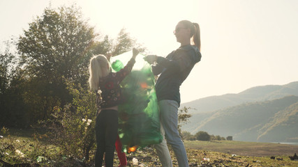 Young mother and her daughter trying to lift up green bag full of trash