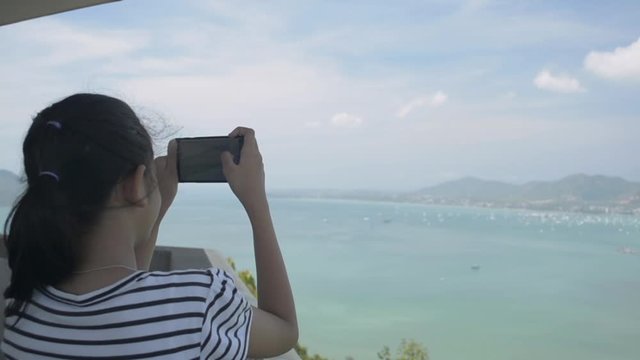 Rear view of pretty girl holding mobile phone while taking picture on view point tower with beautiful scenery of the bay with cloudy blue sky in phuket.