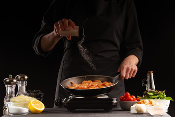 Chef fries seafood, shrimp, sprinkled with seasonings in a pan, frozen in motion. Against the background of vegetables. Black background, cooking and recipe book, cooking and healthy eating.