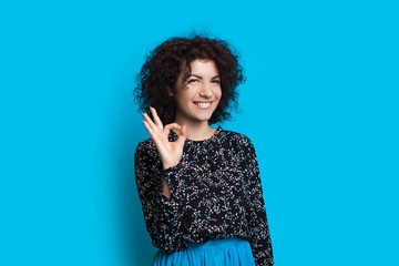 Curly haired caucasian lady is gesturing the okay sign while smiling and posing on a blue background