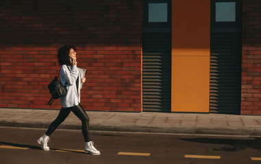 Walking caucasian student with curly hair is speaking on phone while holding a laptop and a bag