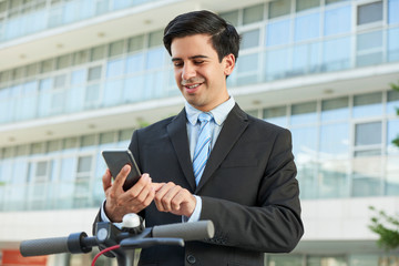 Portrait of young positive entrepreneur checking messages and notifications in his smartphone when walking in the street