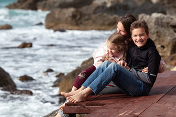 Fototapeta na wymiar Happy mom, eldest son and little daughter sit barefoot on a wooden pier and laugh on the background of the sea and rocks,