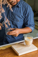 Male barista making froth milk in hot latte coffee in drinking glass at the wooden counter bar. Served on wooden plate.
