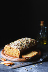 Homemade Pumpkin Bread and a Piece of Bread on a Wooden Plate with a Bottle of Olive Oil in the Background and a Knife and Some Pumpkin Peel Lying Around On the Black Background