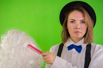 Serious girl in a hat, white shirt, suspenders and bow tie combs a wig with hair on a green...