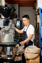 Positive Asian young man operating big machine when roasting coffee at manufacture