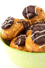 Profiteroles, coated in chocolate, stacked in a pattermed green and white bowl dish.  On a white wood background