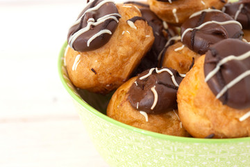 Profiteroles, coated in chocolate, stacked in a pattermed green and white bowl dish.  On a white wood background