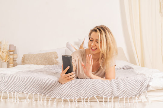 A girl in pajamas is lying on a bed in her apartment with a phone. She is surfing in social networks and smiling. She waving with her hand. Soft pink creamy colors of the picture. Horizontal picture