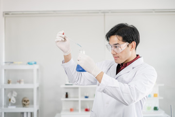 A male scientist with black hair wearing white coat and protective glassware dropping liquid solution from a pipet into a flask in a laboratory setting.
