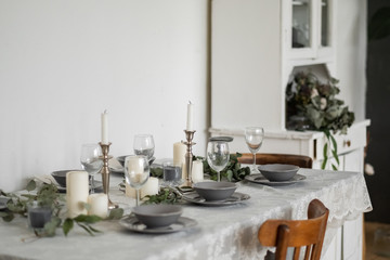 Table setting: gray dishes, white candles, candlesticks, glass goblets, sprigs of eucalyptus on a wooden table 