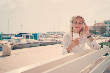 Fototapeta na wymiar Enjoying the sound. Happy young woman with earphones listening music on smartphone while sitting on city embankment sea promenade.