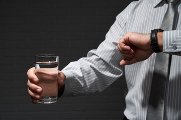 Businessman closeup portrait, he standing and posing with glass of water, looking at wrist watch, dark wall background