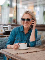 young beautiful blonde woman on a summer morning in sunglasses communicates on a mobile phone in a cafe over a Cup of cappuccino coffee. .