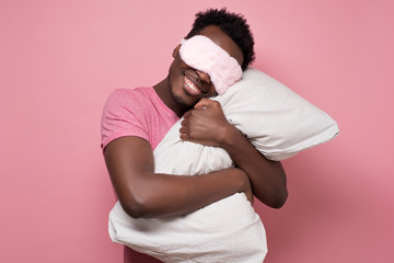 Young african american black man sleeping hugging a pillow wearing sleeping mask. Studio shot on pink wall.