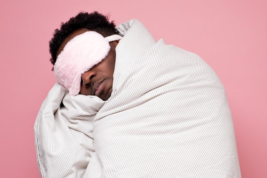 Young african american black man sleeping hugging a pillow wearing sleeping mask. Studio shot on pink wall.