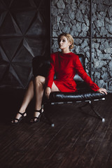 Beautiful woman lady in a red dress sits sadly on a leather chair in a loft style. Soft selective focus. Beauty, fashion.