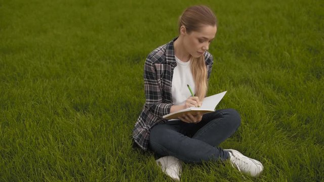 teen girl sits on the grass and takes notes in a notebook. teen girl sits on the grass and takes notes in a notebook. 4K
