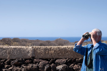One senior man with binoculars  - beard and white hair - walking outdoor in an arid landscape with cactus - blue sea and sunny day