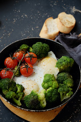 Healthy breakfast: broccoli, cherry tomatoes and fried eggs in a pan, which stands on a wooden stand on a dark background. 