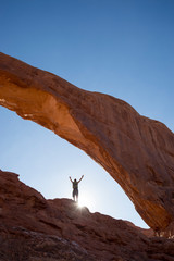 Backlit view of distant hiker raising his arms to the sky standing under a dramatic natural rock arch in front of golden sun