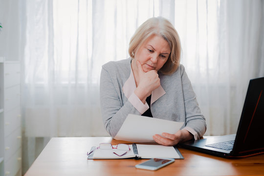 Older Senior Woman Struggles At The Computer With Paper Documents, Puzzled By The Business Decision