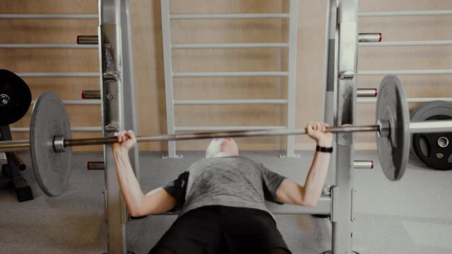 Young white Caucasian man in grey sweater at a gym lifts weights on a bench with ease, then suddenly it becomes really heavy and he fails completely lifting it. He struggles intensively.