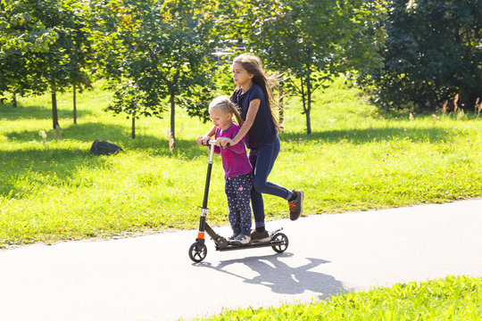 Two happy caucasian kid girls having fun riding on a cick scooter in a city park in summer