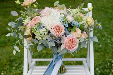 Close up of bridal bouquet of pink roses, blue flowers and greenery on white wood chair outdoors, copy space. Wedding concept