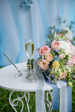 Close up of bridal bouquet of pink and blue flowers, glass of champagne and wedding rings on white wood table outdoors, copy space. Wedding concept