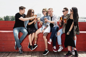 Young happy friends drinking beer before festival at outdoors pub on roof, toasting and laughing, copy space. Friendship and celebration concept