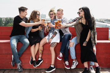 Young happy friends drinking beer before festival at outdoors pub on roof, toasting and laughing, copy space. Friendship and celebration concept