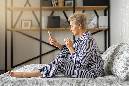 Adult Elderly Woman In Pajamas In Bed In The Bedroom With A Mug And A Phone In Her Hands