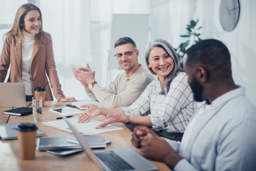 Fototapeta premium smiling asian woman pointing with hands at folder and looking at african american colleague in creative agency