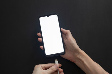 The hand holds a smartphone with the display turned on and the second inserts the charger cable.