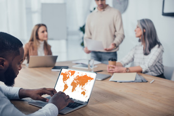 selective focus of african american man using laptop with map and his colleagues talking on background