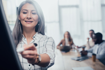 selective focus of smiling asian businesswoman writing on flipchart and her colleagues on background